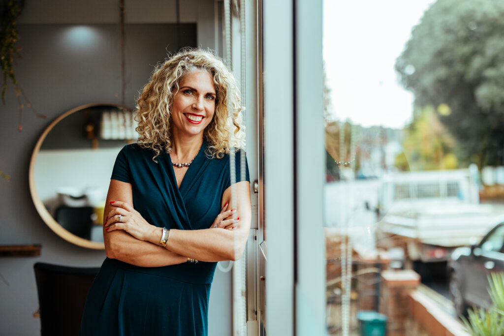 Anna Balcombe standing by the salon window in a teal dress